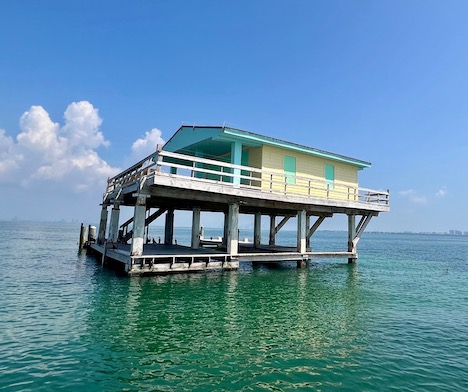 Photo of colorful yellos and green Stiltsville house  from a boat tour on a sunny day with clear blue water and sky in background.