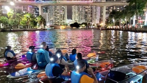 A group on colorful clear kayaks against the night skyline of miami during a nighttime kayak tour of the miami river.