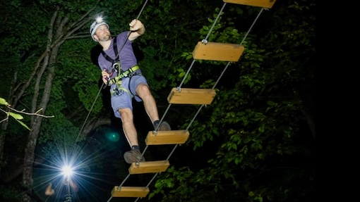 A man is on a treetop trekking zip line experience at night with headlamp on overlooking downtown Miami skyline.